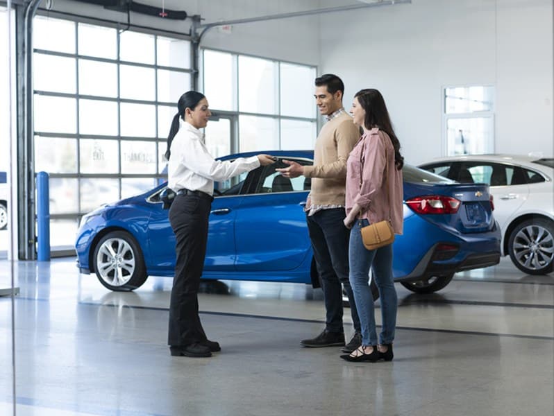 Couple receives car keys from a dealer in a showroom, standing near a blue vehicle.
