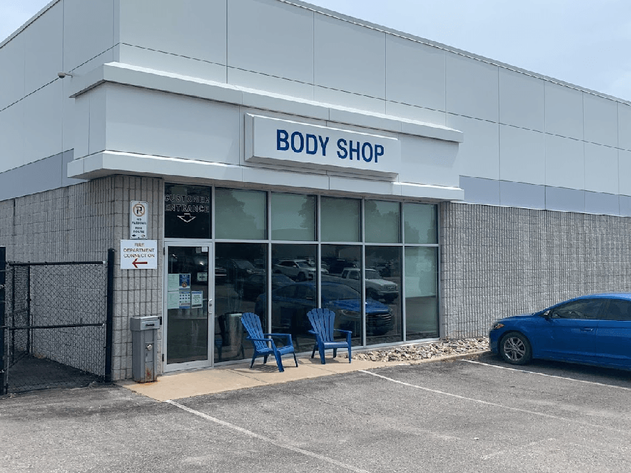 Auto body shop exterior with large windows, blue chairs, and parked car. Vehicle repair services.