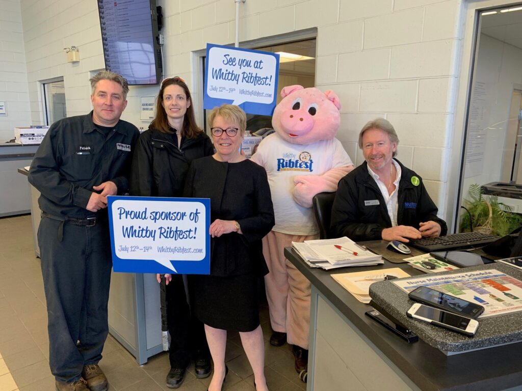 Whitby Ribfest sponsors posing with a pink mascot, showcasing event signage.