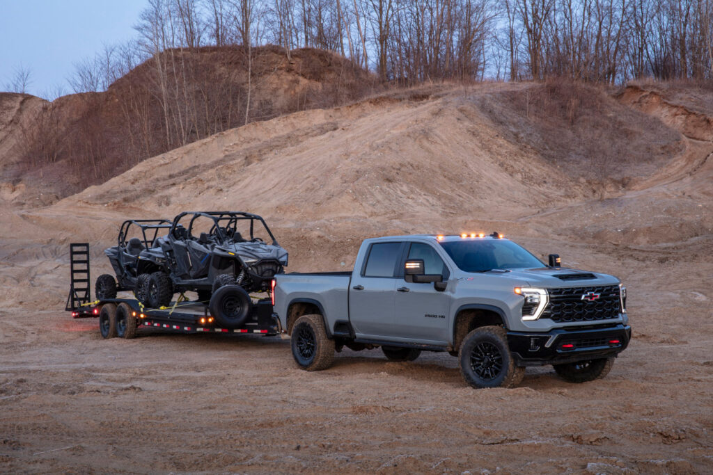 Gray pickup truck towing off-road vehicles on a trailer in a rugged, hilly landscape.