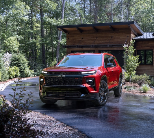 Red SUV parked on a wet driveway in front of a modern wooden house surrounded by trees.