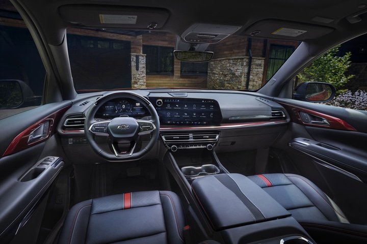 Interior view of a modern car dashboard featuring a multi-function steering wheel, digital display, and touchscreen with various apps. The trim includes red accents on air vents. Urban street scene visible through the windscreen.