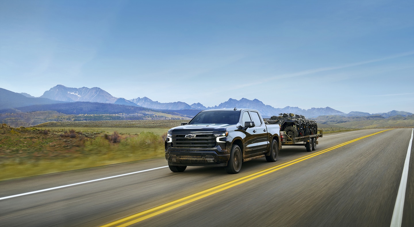 Black pickup truck towing a trailer on a scenic mountain road under a clear blue sky.