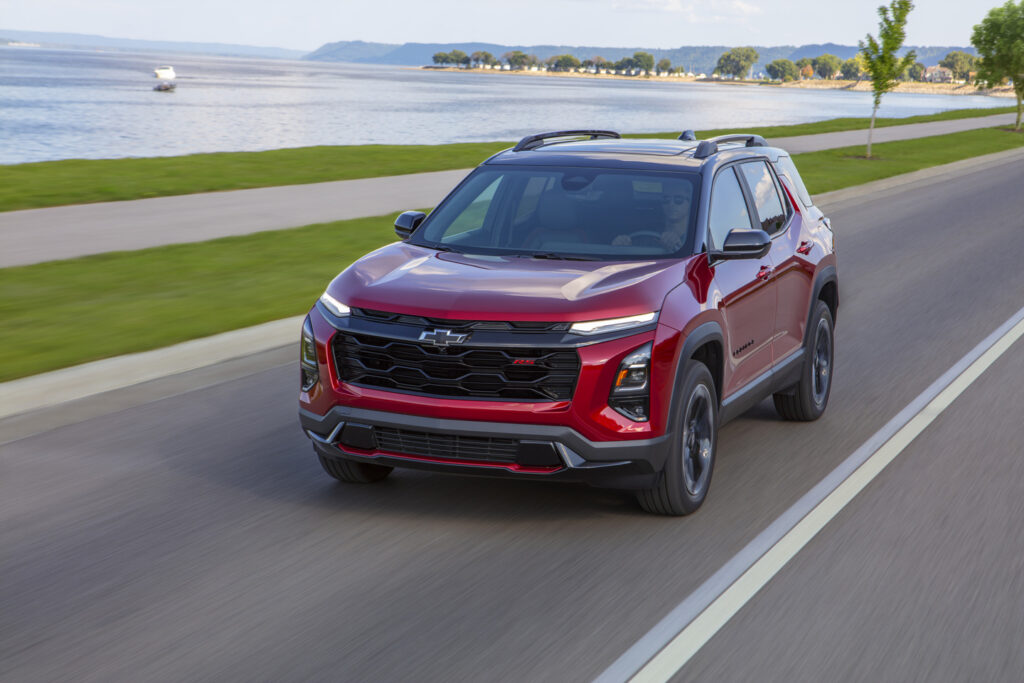 Red Chevrolet SUV driving along a scenic lakeside road, with hills in the background.