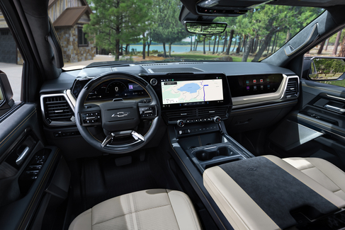 View from the backseat of a car showing two people in the front seats. The driver is holding the steering wheel. The sunroof is open, revealing a blue sky and tall buildings outside. The dashboard and rearview mirror are visible.