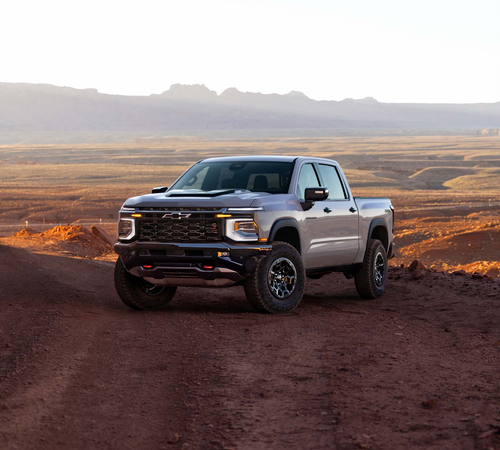 Silver pickup truck on a rugged dirt road in a desert landscape at sunrise.