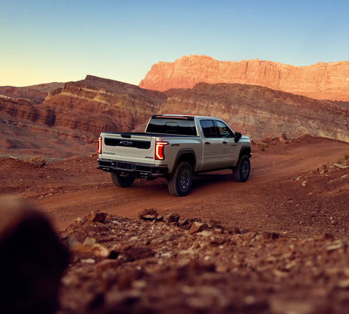 Truck driving on a desert road with rocky landscape and sunset-lit cliffs in the background.