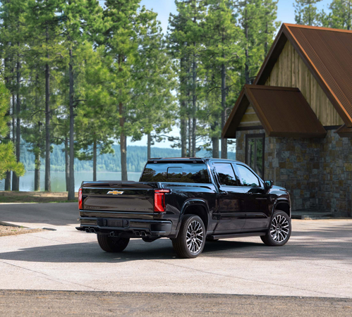 Black truck parked in front of a cabin, surrounded by tall trees and a serene lake view.