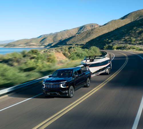 Black pickup truck towing a boat on a scenic mountain road by a lake under clear blue skies.