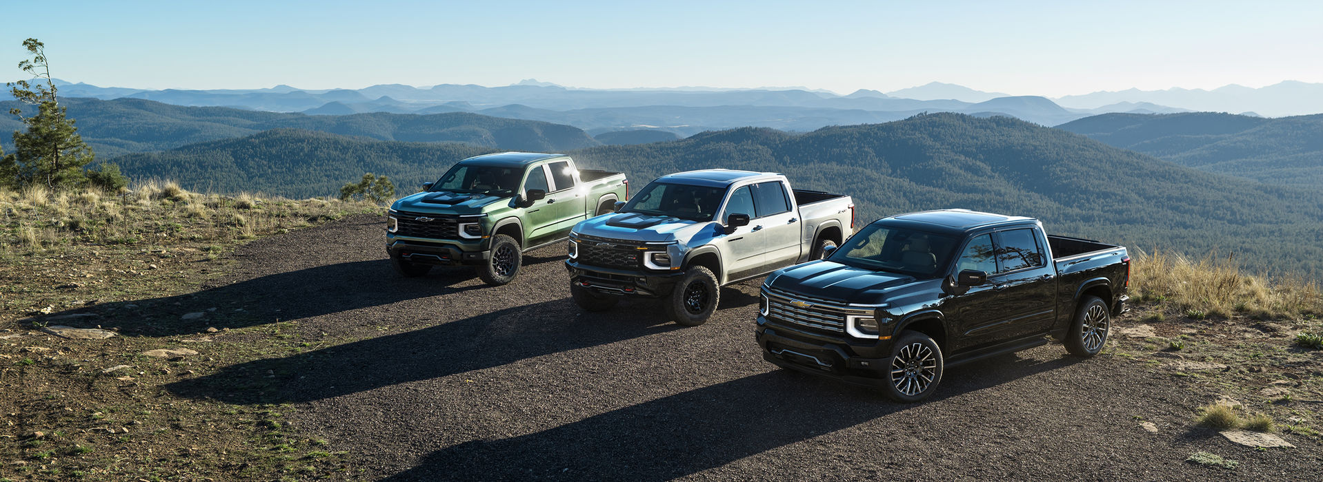 Three 2027 Chevrolet Silverado 1500 trucks on a hilltop above forested mountains