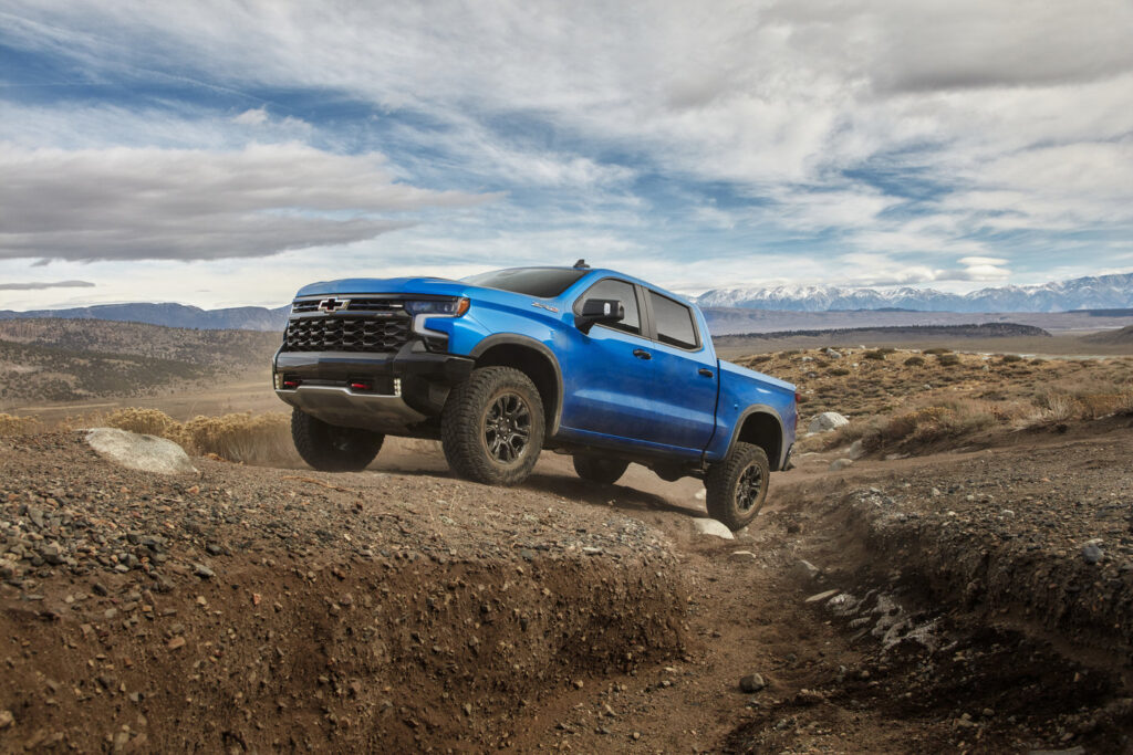 Blue pickup truck navigating rocky terrain with mountains in the background under a cloudy sky.