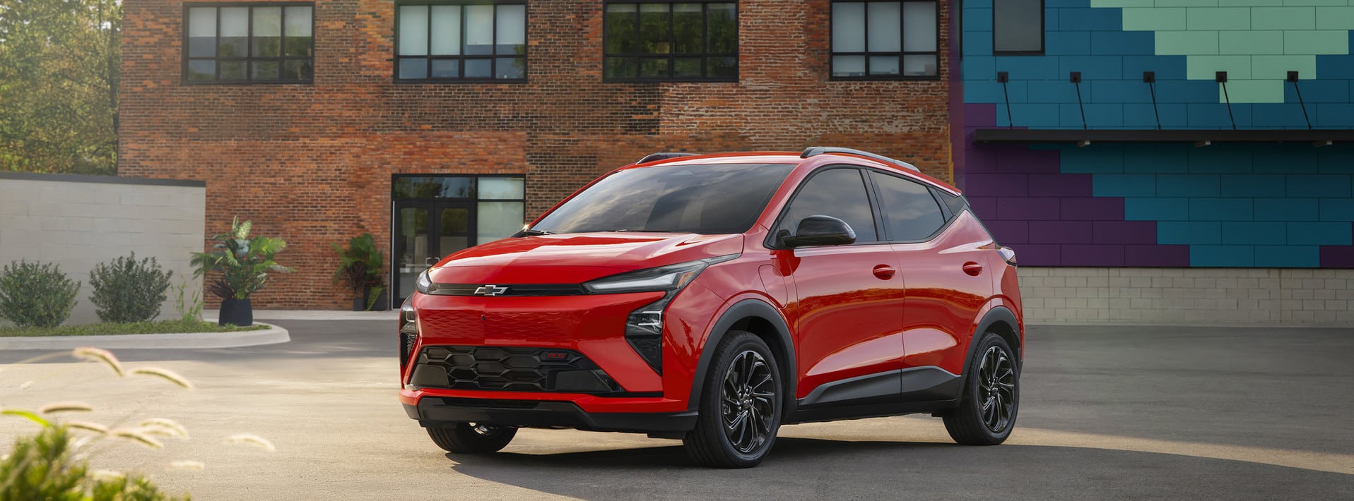 A red SUV with black and chrome accents is driving on a city street. Its a four-door vehicle with large alloy wheels, a sleek design, and tinted windows. The background features blurred buildings and trees.