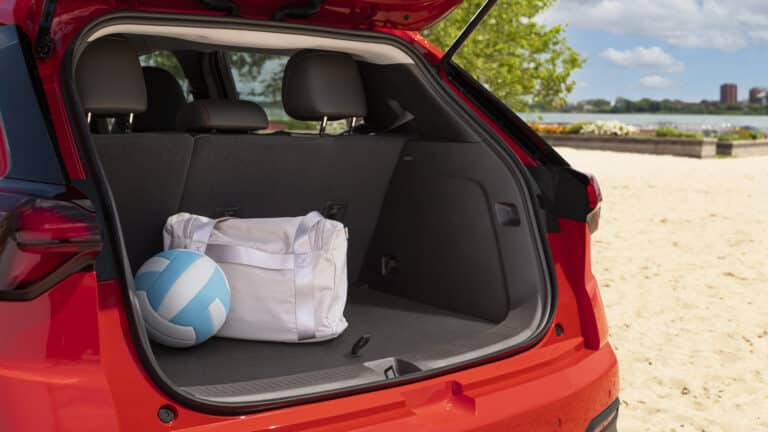 Open trunk of a red car at a beach, containing a white bag and blue volleyball.