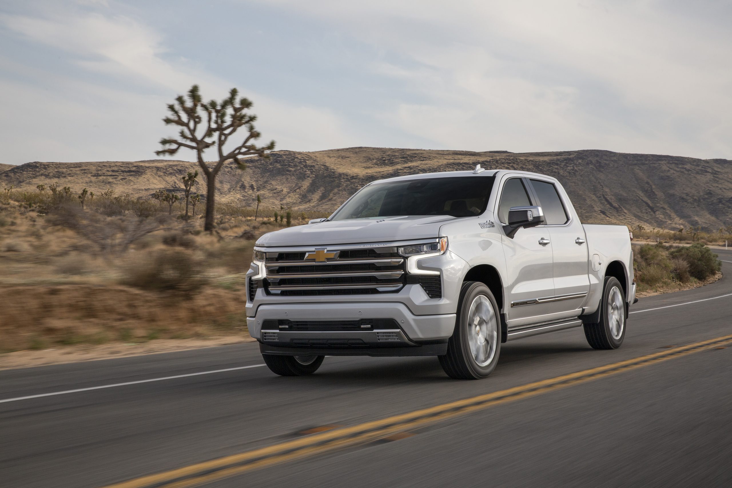 White pickup truck driving on a scenic desert road with hills and a Joshua tree in the background.