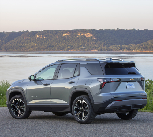 Gray SUV parked by a lake with a wooded hill in the background.