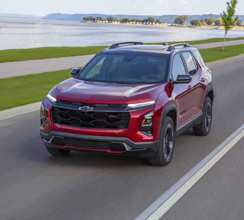 Red Chevrolet SUV driving along a lakeside road, with trees and distant hills in the background.
