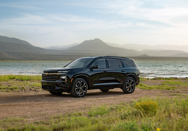 A red SUV with black and chrome accents is driving on a city street. Its a four-door vehicle with large alloy wheels, a sleek design, and tinted windows. The background features blurred buildings and trees.
