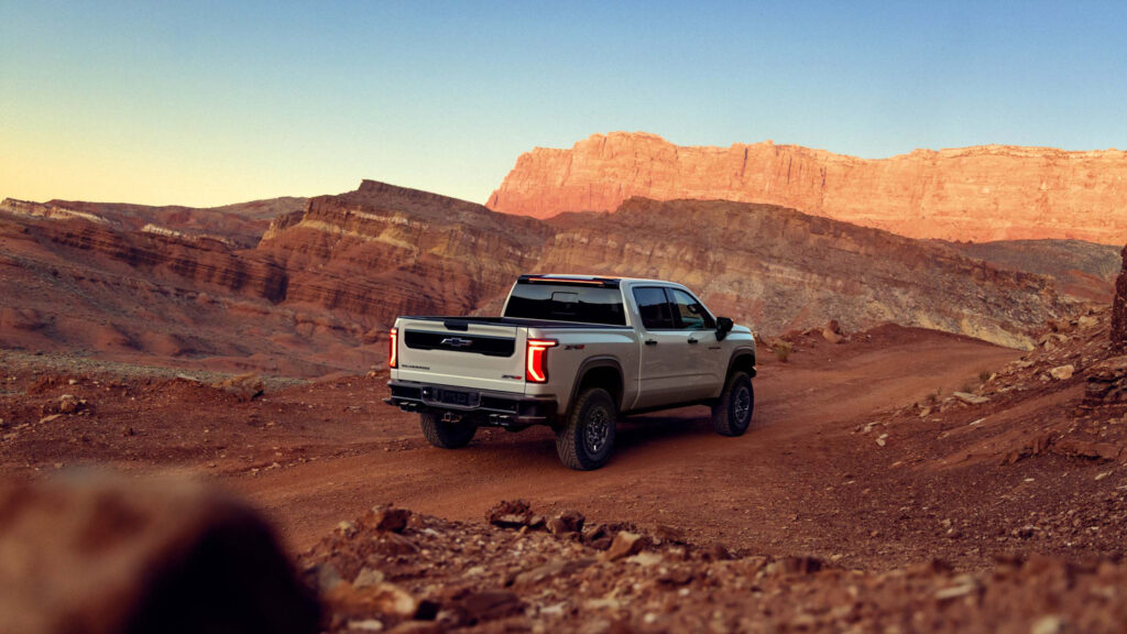 A white pickup truck drives on a rugged dirt road in a canyon landscape at sunset.